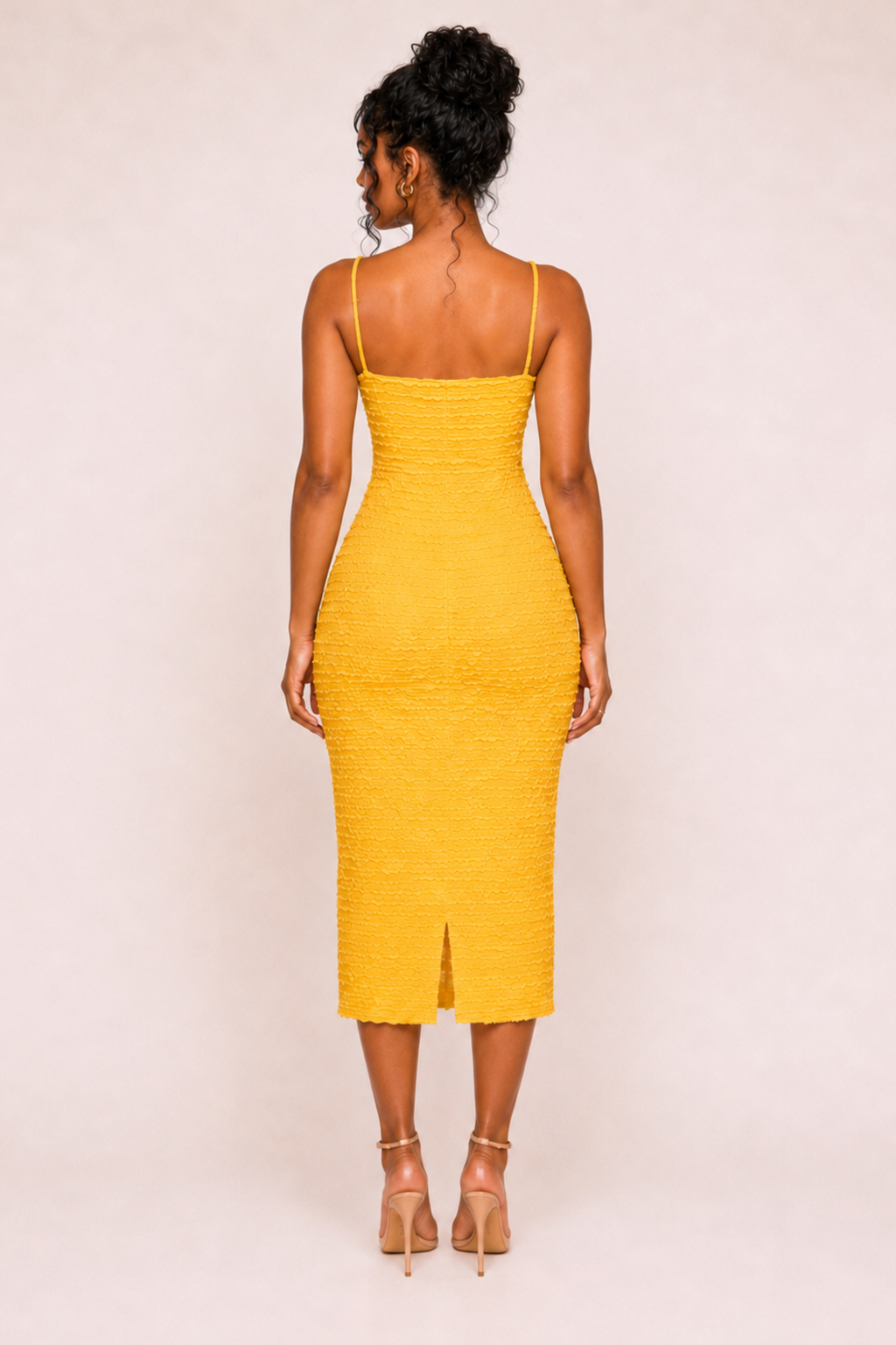 A woman viewed from the back wearing a bright yellow textured spaghetti-strap midi dress with a front slit, paired with nude high-heeled sandals, against a plain light beige studio background.