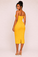 A woman modeling a bright yellow textured midi dress with thin straps and a side slit, shown from the back against a plain light beige studio background.
