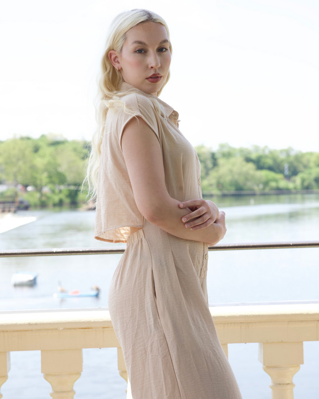 Woman in a beige dress standing by a waterfront with trees in the background