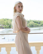 Woman in a beige dress standing by a waterfront with trees in the background