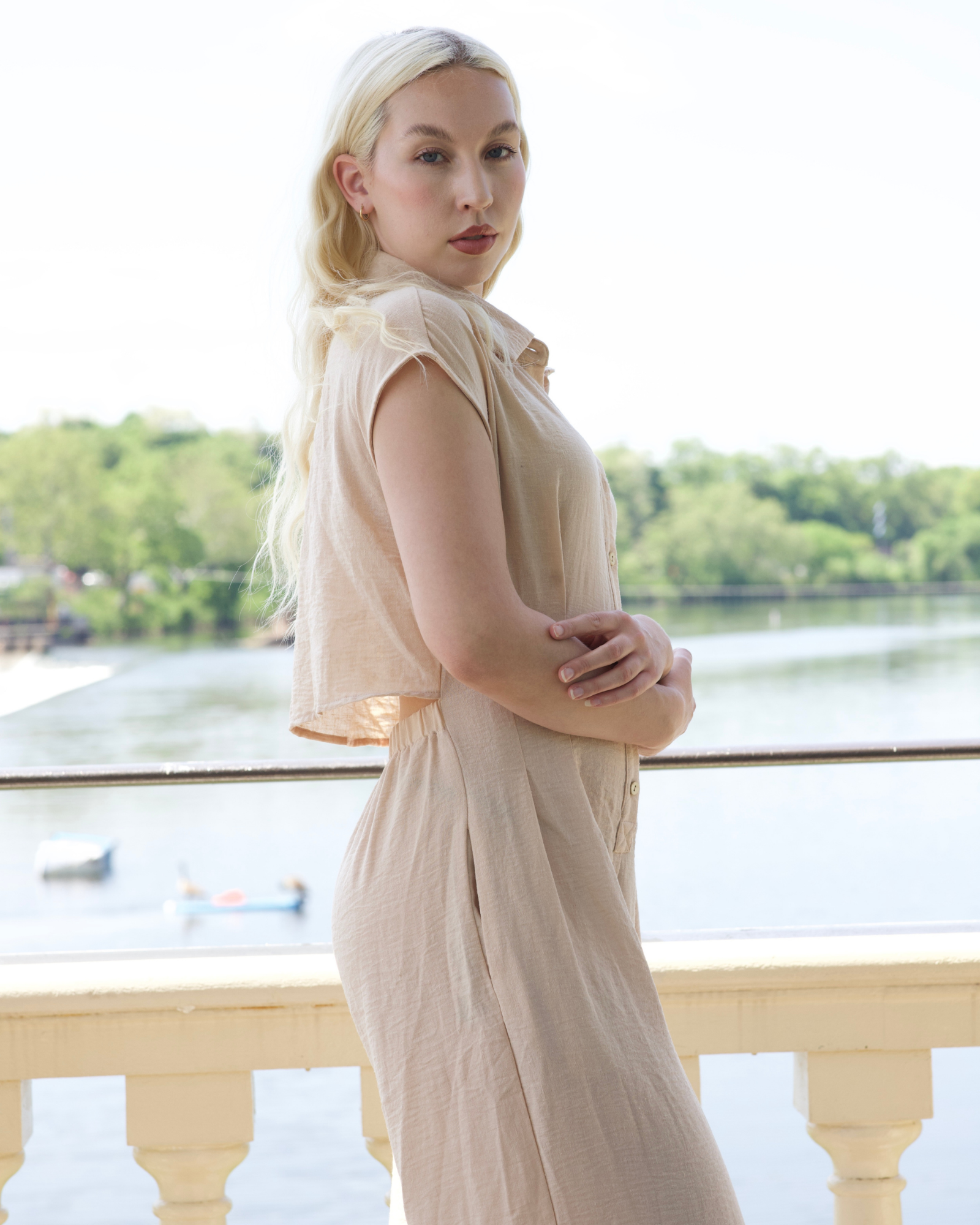 Woman in a beige dress standing by a waterfront with trees in the background