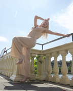 Woman in a beige dress posing on a balcony with a clear blue sky and water in the background
